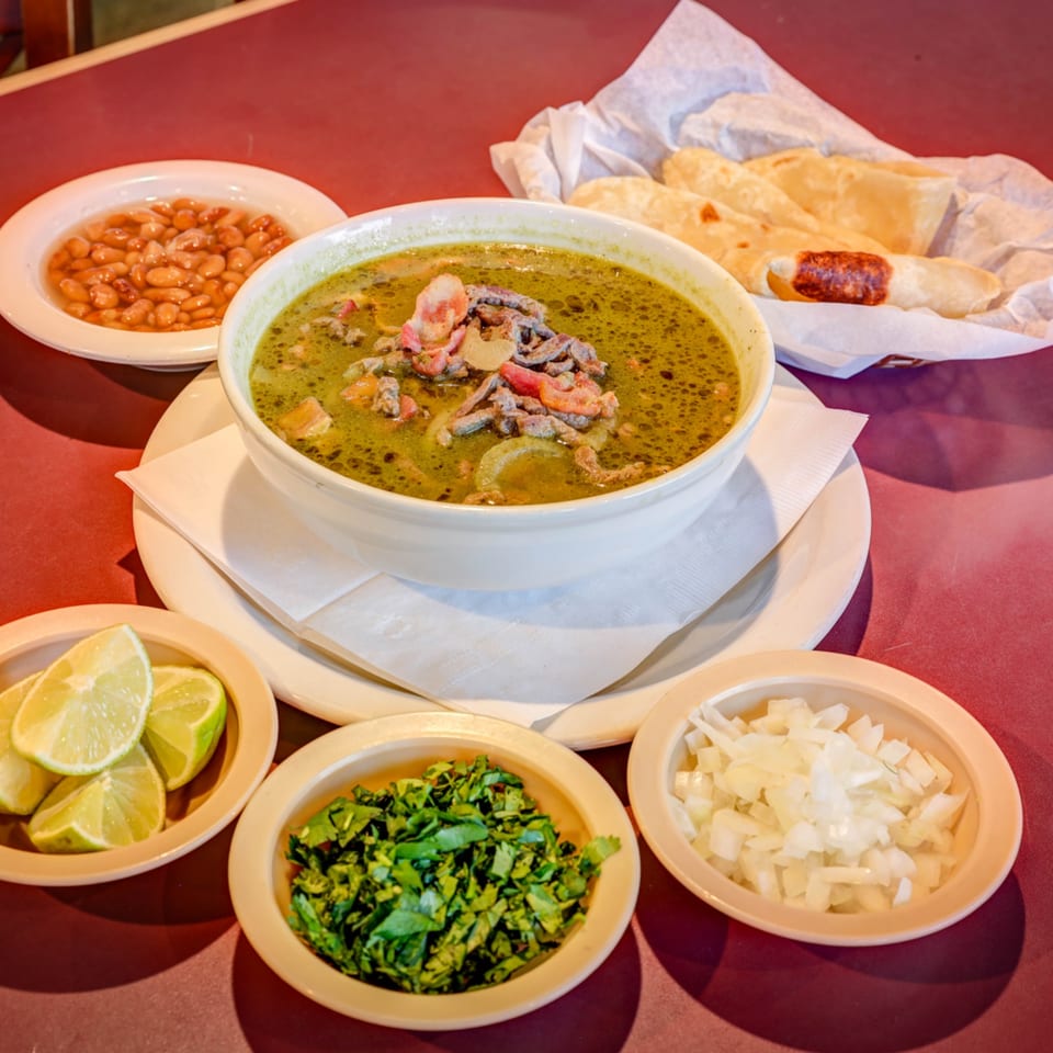 A bowl of green Mexican soup with meat, beans, tortillas, limes, cilantro, and onions.