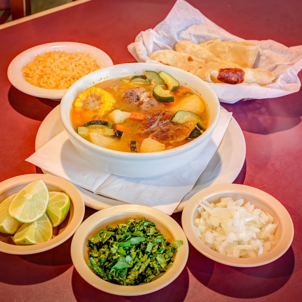 A bowl of Mexican beef soup with corn, zucchini, and carrots, served with rice, tortillas, and garnishes.